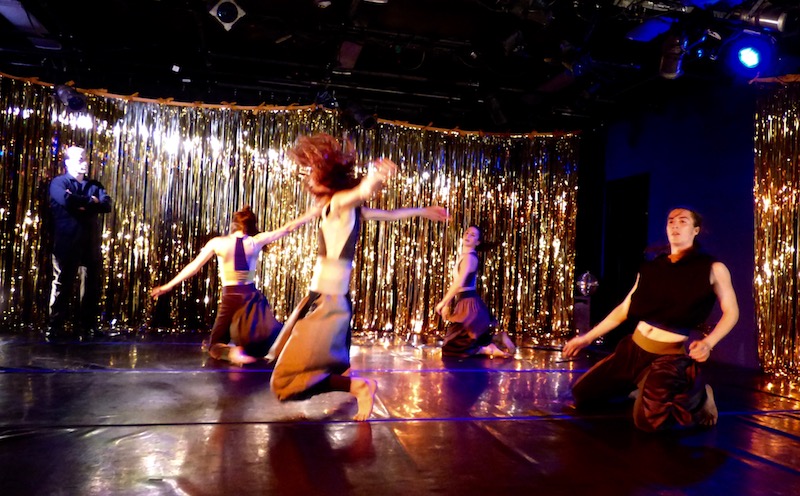 A group of dancers kneel on stage. Around the perimeter of the room are gold streamers. A man with his arms crossed stands and watches them.